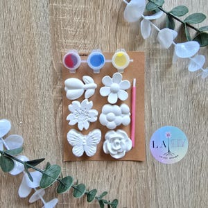 Op de afbeelding: Een knutselset met witte gipsen bloem- en vlindervormen, verfpotjes met rode, blauwe en gele verf en een roze penseel. De set bevat een ronde sticker met de tekst "LA ELY ATELIER".