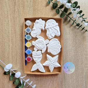 Pode incluir: Um kit de pintura de criaturas marinhas para pintar, com um cavalo-marinho, uma água-viva, uma tartaruga, uma concha, uma sereia e uma estrela-do-mar. Inclui potes de tinta em várias cores e dois pincéis. O kit é apresentado numa caixa de cartão castanha.