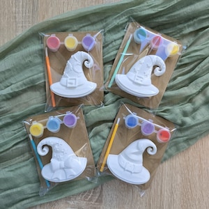 Pode incluir: Quatro kits de artesanato individuais, cada um contendo uma figura de chapéu de bruxa de cerâmica branca, um pincel pequeno e três potes de tinta pequenos com várias cores. Cada kit está selado numa embalagem de plástico transparente.
