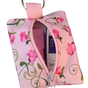 以下が含まれることがあります： ピンクのジッパー付きポーチで、ピンクのバラと緑のつるが花柄になっています。ポーチにはシルバーのキーリングとシルバーのジッパーが付いています。ポーチが開いていて、ピンクと白のUSBドライブが入っています。