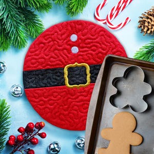 May include: A red quilted Santa Claus-themed trivet with a black belt and gold buckle, next to a baking sheet with a gingerbread man cookie cutter and a baked gingerbread man. Christmas and holiday theme.