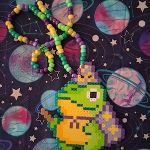 May include: A colorful beaded necklace with a pendant featuring a pixelated green and yellow dinosaur character. The necklace has a beaded chain in shades of green, yellow, and purple. The background is a dark blue fabric with a space-themed pattern.