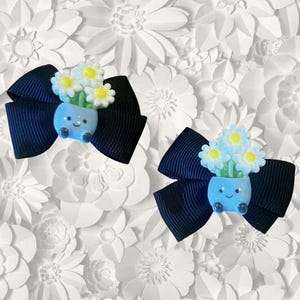 May include: Two navy blue hair bows with a light blue flower pot design. Each bow features a smiling flower pot with white and yellow flowers. The bows are set against a white floral background.
