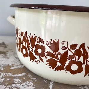 May include: White enamel pot with a brown floral design around the rim. The pot has a brown rim and two handles.