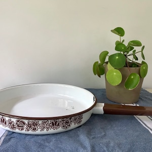 May include: White enamel frying pan with a brown floral design around the rim and a brown handle. The pan is sitting on a blue and white striped towel.