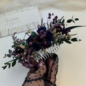 Op de afbeelding: Een bloemenhaarkam met dieppaarse en marineblauwe bloemen, geaccentueerd met groene bladeren en takjes. De kam wordt vastgehouden door een hand met een zwarte handschoen met stippen. Een decoratief haaraccessoire.