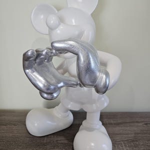 May include: A white Mickey Mouse figurine with silver-colored hands. The figurine is standing on a wooden surface. The figure is a stylized representation of the iconic cartoon character.