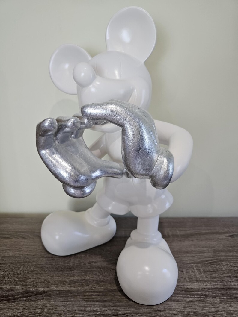 May include: A white Mickey Mouse figurine with silver-colored hands. The figurine is standing on a wooden surface. The figure is a stylized representation of the iconic cartoon character.