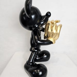May include: A glossy black Mickey Mouse figurine holding a gold-colored hand. The figurine has large, rounded ears and a classic cartoon design. The gold hand appears to be holding a cluster of gold objects. The figurine is standing on a white surface.