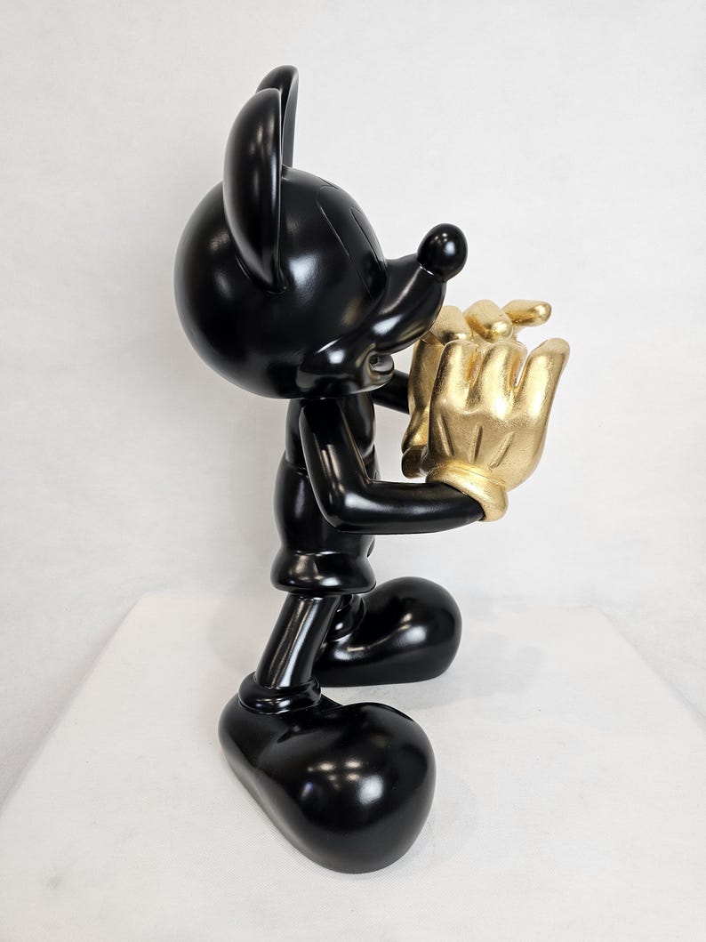 May include: A glossy black Mickey Mouse figurine holding a gold-colored hand. The figurine has large, rounded ears and a classic cartoon design. The gold hand appears to be holding a cluster of gold objects. The figurine is standing on a white surface.
