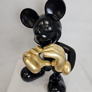May include: A glossy black Mickey Mouse figurine with gold-colored hands. The sculpture features Mickey in a classic pose, with his hands clasped together. The figurine is set against a plain white background.