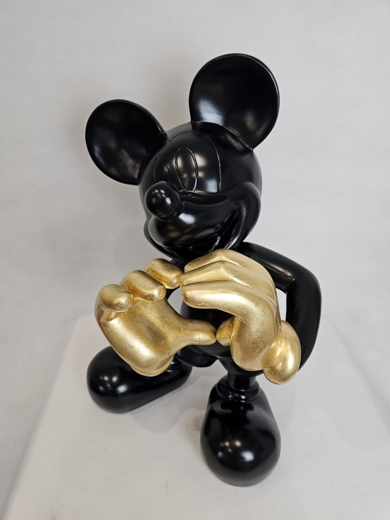 May include: A glossy black Mickey Mouse figurine with gold-colored hands. The sculpture features Mickey in a classic pose, with his hands clasped together. The figurine is set against a plain white background.