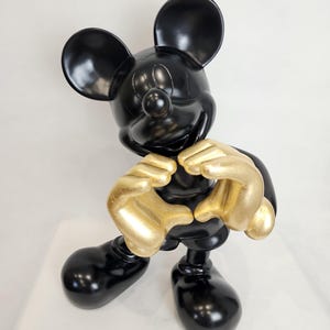 May include: A black Mickey Mouse figurine with gold-colored hands forming a heart shape. The figurine has large black ears, a round nose, and large black feet. The figure is standing on a white surface.