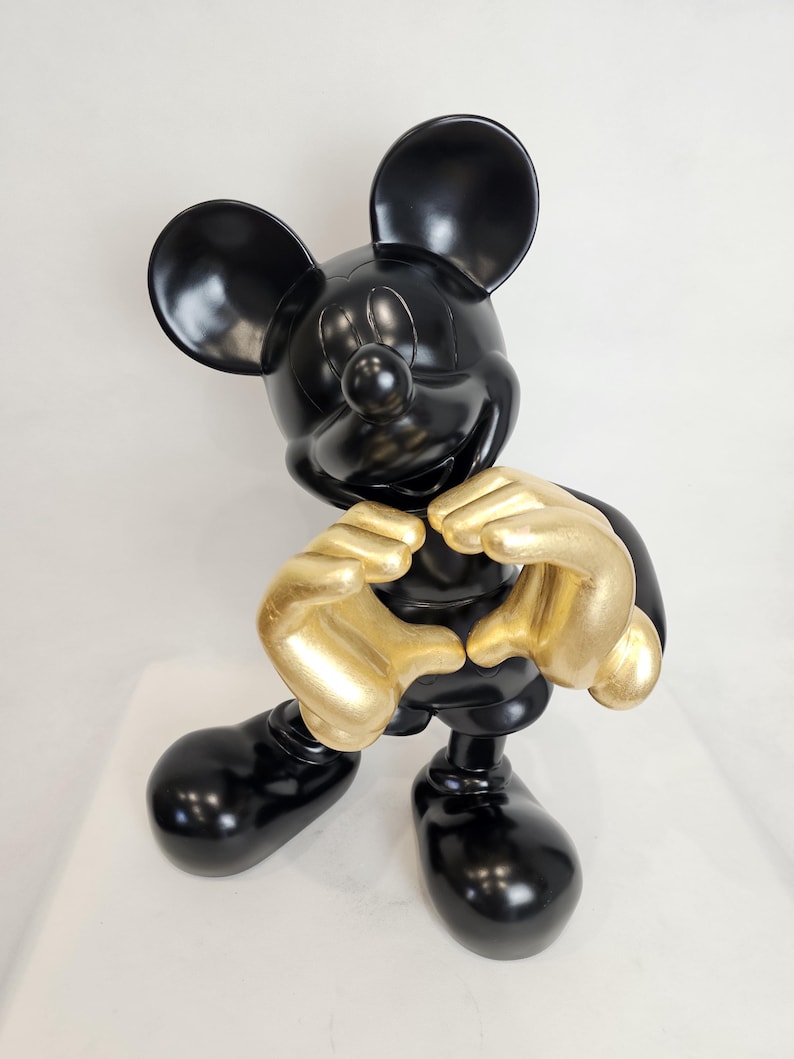 May include: A black Mickey Mouse figurine with gold-colored hands forming a heart shape. The figurine has large black ears, a round nose, and large black feet. The figure is standing on a white surface.