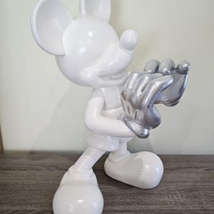 May include: A white Mickey Mouse figurine with silver-colored hands. The figure is standing on a wooden surface. The figurine has large, round ears and a smiling expression.