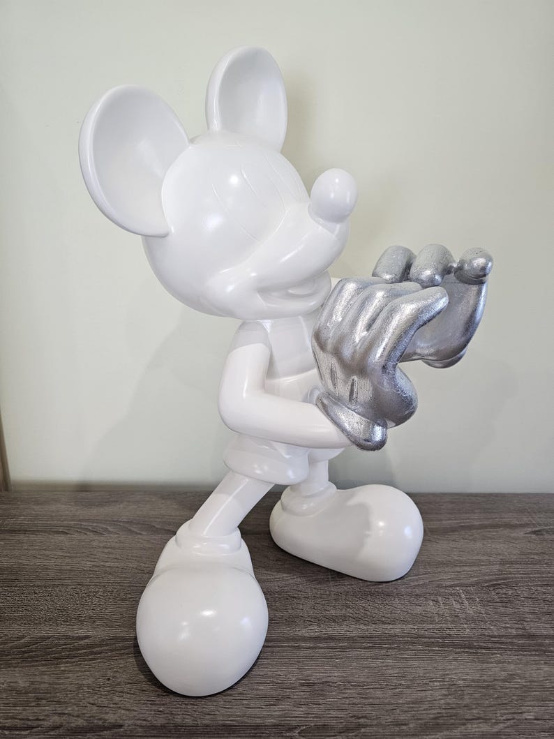 May include: A white Mickey Mouse figurine with silver-colored hands. The figure is standing on a wooden surface. The figurine has large, round ears and a smiling expression.