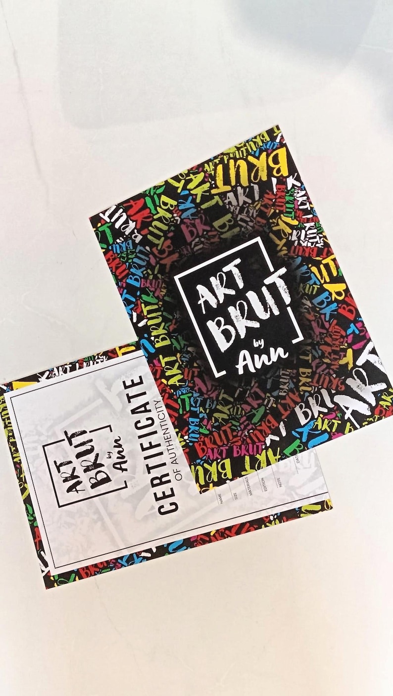 May include: Two rectangular art prints and a certificate of authenticity. The art print has a black background with colorful text that says "ART BRUT" in a graffiti style. The certificate is white with the words "ART BRUT by Ann".