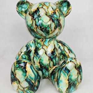 May include: A decorative teddy bear figurine with a marbled design in shades of green, teal, and gold. The bear is sitting with its arms and legs outstretched. The figurine has a glossy finish.