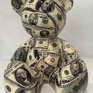May include: A decorative bear figurine covered in a collage of United States currency prints. The surface features various denominations including one, five, fifty, and one-hundred dollar bills with portraits of historical figures like Benjamin Franklin.