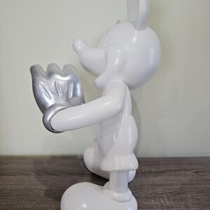 May include: A white Mickey Mouse figurine with a silver-colored glove. The figurine is in a standing pose, with one hand raised. The figure is on a wooden surface, with a light green wall in the background. The figurine is a collectible item.