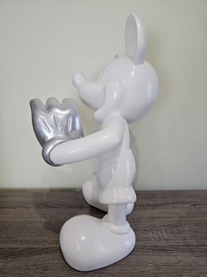 May include: A white Mickey Mouse figurine with a silver-colored glove. The figurine is in a standing pose, with one hand raised. The figure is on a wooden surface, with a light green wall in the background. The figurine is a collectible item.