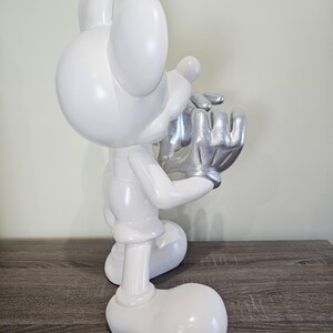 May include: A white figurine of a cartoon mouse with silver hands and a silver face. The mouse is in a kneeling position, holding its hands up to its face. The figurine is on a wooden surface.