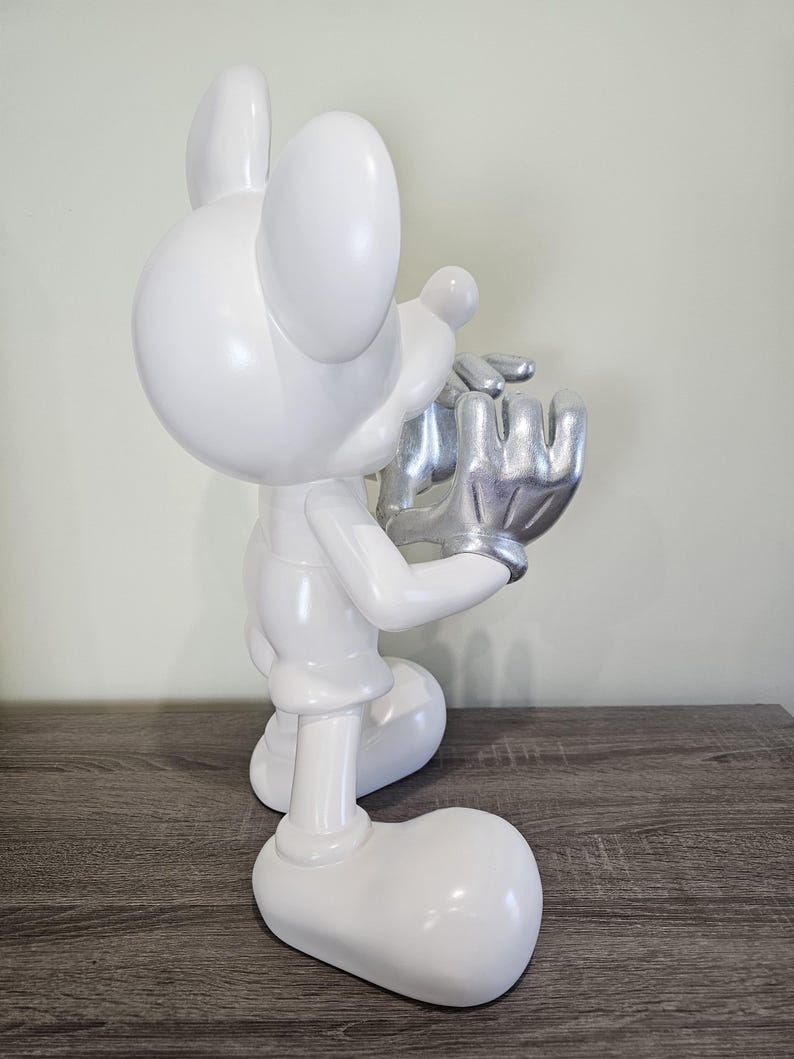 May include: A white figurine of a cartoon mouse with silver hands and a silver face. The mouse is in a kneeling position, holding its hands up to its face. The figurine is on a wooden surface.