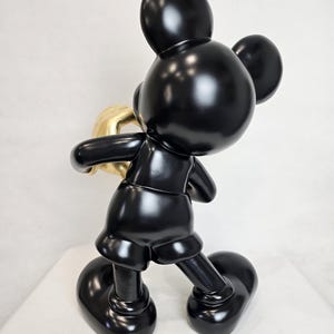 May include: A glossy black Mickey Mouse sculpture holding a gold-colored object. The figure is posed in a walking stance, with large, rounded feet and ears. The sculpture has a smooth, reflective surface.