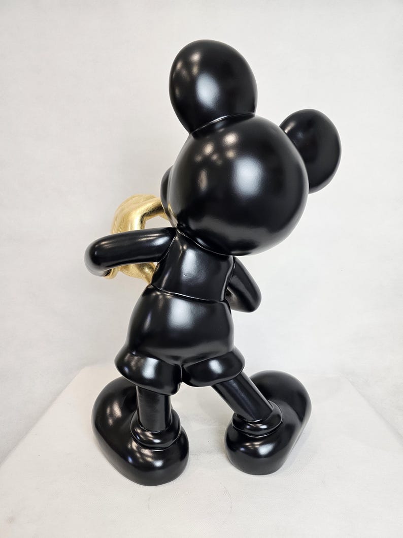 May include: A glossy black Mickey Mouse sculpture holding a gold-colored object. The figure is posed in a walking stance, with large, rounded feet and ears. The sculpture has a smooth, reflective surface.