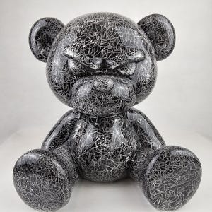 Op de afbeelding: Een zwart en zilver decoratief teddybeerbeeldje. De beer zit in een zittende positie met uitgestrekte armen en benen. Het oppervlak is bedekt met een zilverkleurig, webachtig patroon. De beer heeft een glanzende afwerking.
