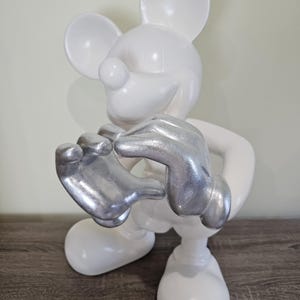 May include: A white Mickey Mouse figurine with silver-colored hands. The figurine is in a standing pose, with its hands clasped together. The figure has large, round ears and a smiling expression. The figurine is on a wooden surface.