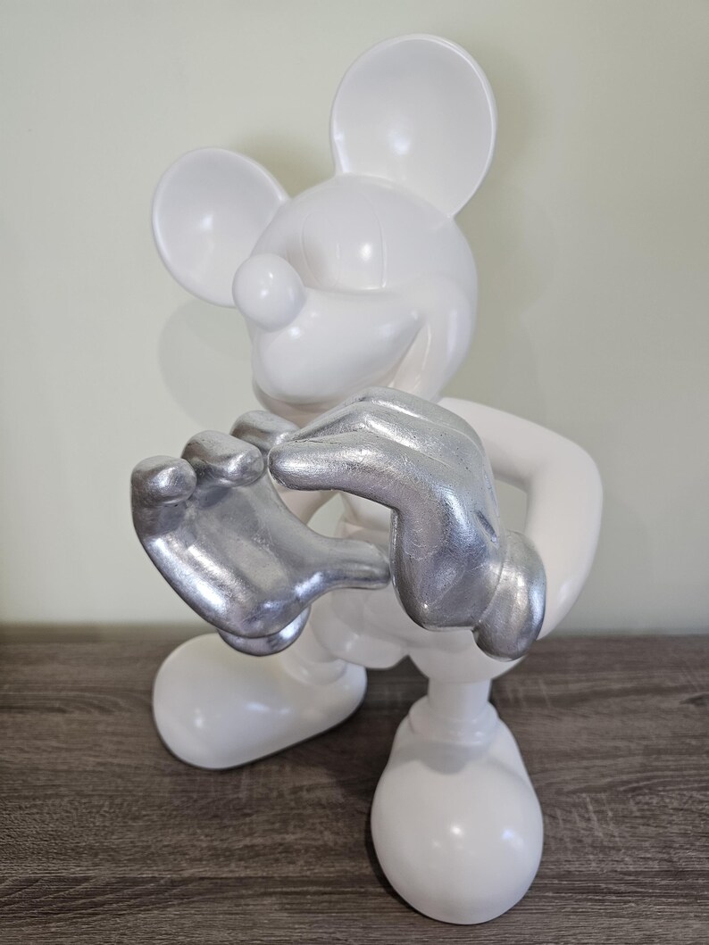 May include: A white Mickey Mouse figurine with silver-colored hands. The figurine is in a standing pose, with its hands clasped together. The figure has large, round ears and a smiling expression. The figurine is on a wooden surface.