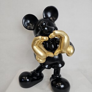 May include: A glossy black Mickey Mouse figurine with gold-colored hands forming a heart shape. The sculpture has large, rounded ears and feet, and is set against a plain white background. The figurine is a decorative item.