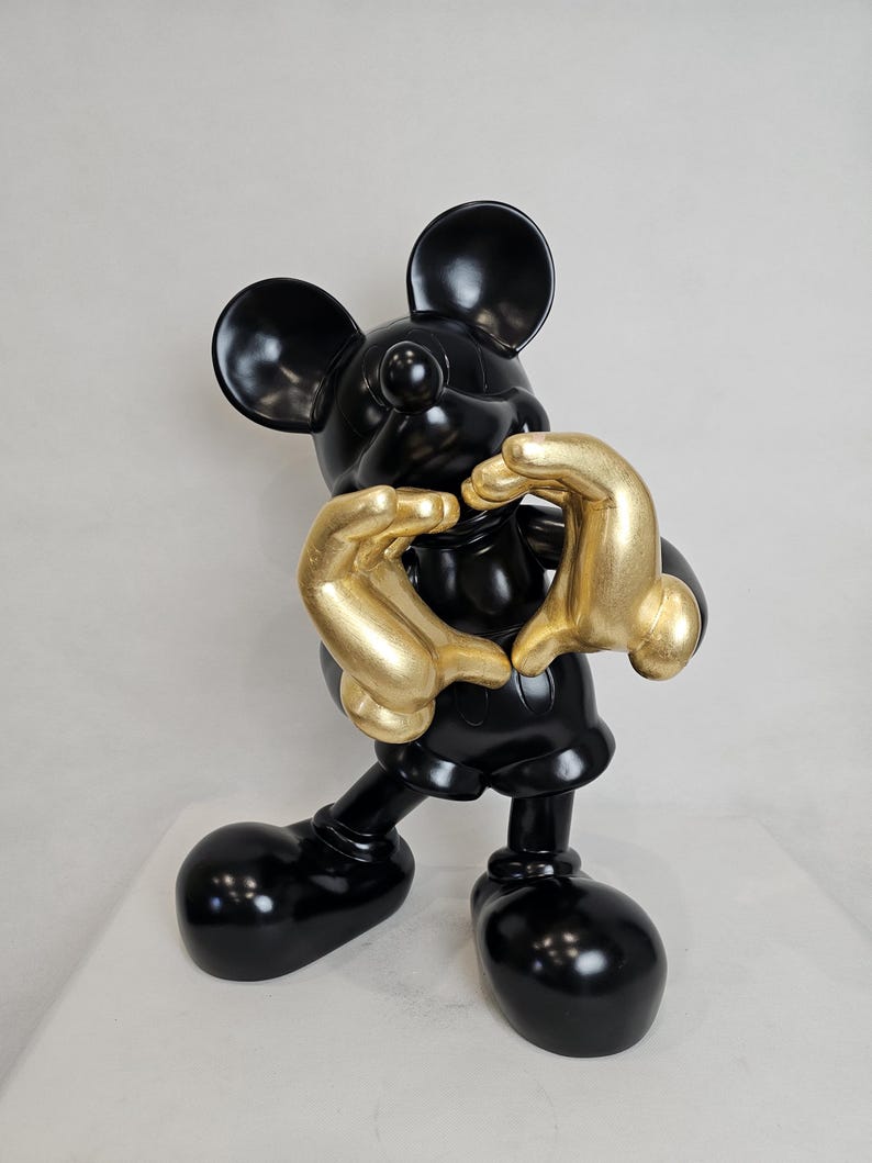 May include: A glossy black Mickey Mouse figurine with gold-colored hands forming a heart shape. The sculpture has large, rounded ears and feet, and is set against a plain white background. The figurine is a decorative item.