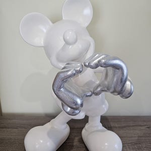 May include: A white Mickey Mouse figurine with silver hands. The figurine is posed with its hands forming a heart shape. The figure is standing on a wooden surface.