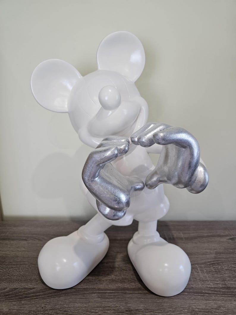 May include: A white Mickey Mouse figurine with silver hands. The figurine is posed with its hands forming a heart shape. The figure is standing on a wooden surface.