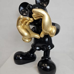 May include: A glossy black Mickey Mouse figurine with gold-colored hands. The figure is in a playful pose, with its hands covering its face. The figurine has large, round black ears and feet.