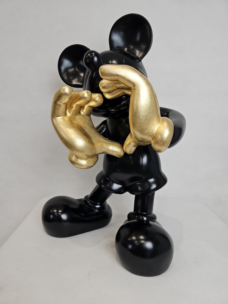 May include: A glossy black Mickey Mouse figurine with gold-colored hands. The figure is in a playful pose, with its hands covering its face. The figurine has large, round black ears and feet.