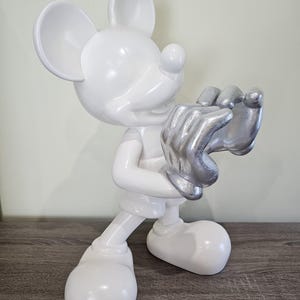 May include: A white Mickey Mouse figurine with silver-colored hands. The figurine is standing on a wooden surface. The figure is a glossy white color with large ears and a smiling face.