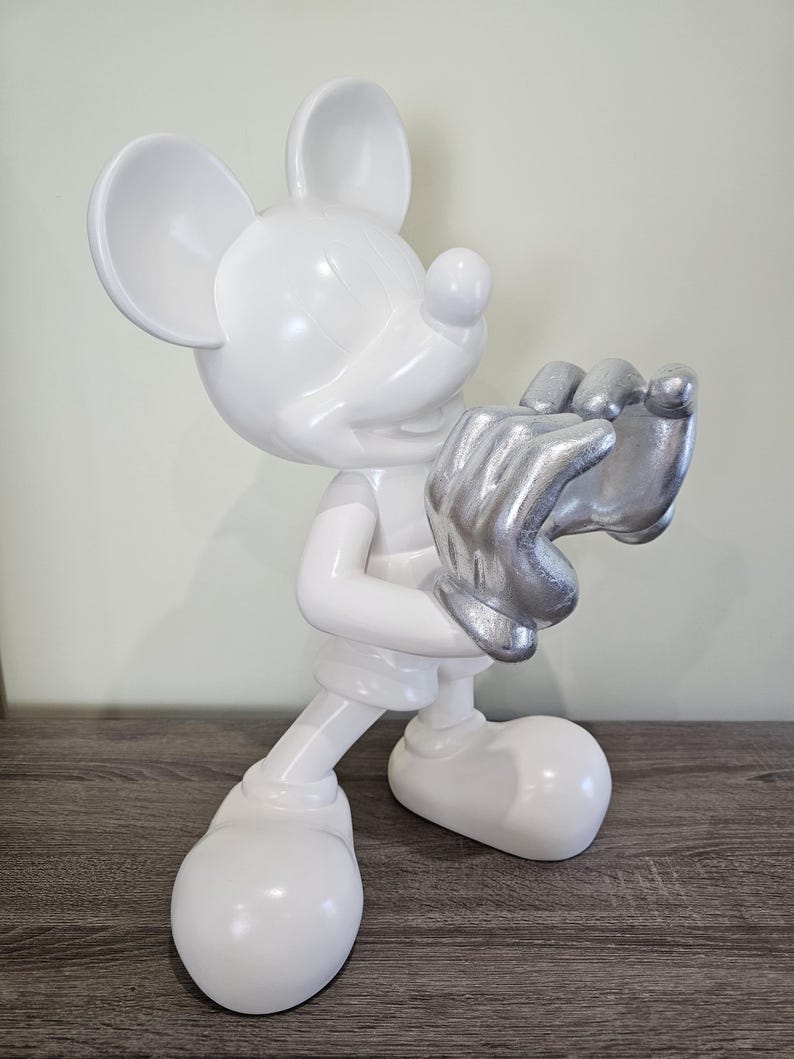 May include: A white Mickey Mouse figurine with silver-colored hands. The figurine is standing on a wooden surface. The figure is a glossy white color with large ears and a smiling face.