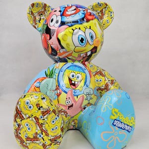 Op de afbeelding: Een kleurrijk berenbeeldje met personages uit de animatieserie SpongeBob SquarePants. Het lichaam van de beer is bedekt met afbeeldingen van SpongeBob, Patrick Star, Octo Tentakel en andere personages. Op de rechterpoot van de beer staat "SpongeBob SquarePants".