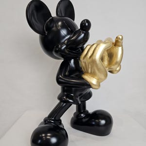 May include: A glossy black Mickey Mouse figurine holding golden hands. The sculpture features Mickey's iconic large ears, round feet, and a cheerful expression. The golden hands add a striking contrast to the black figure.