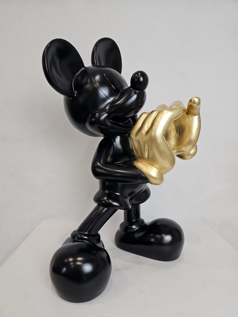 May include: A glossy black Mickey Mouse figurine holding golden hands. The sculpture features Mickey's iconic large ears, round feet, and a cheerful expression. The golden hands add a striking contrast to the black figure.
