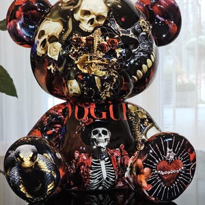 Può includere: Statuetta decorativa a forma di orso con finitura lucida. L'orso è decorato con un collage di teschi, rose, serpenti e la scritta "VOGUE". La tavolozza dei colori comprende nero, rosso, bianco e oro.