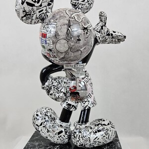 Pode incluir: Uma figura de Mickey Mouse em preto e branco, coberta de recortes de jornais e texto. A figura est&aacute; em p&eacute; sobre uma base de m&aacute;rmore preto e branco.