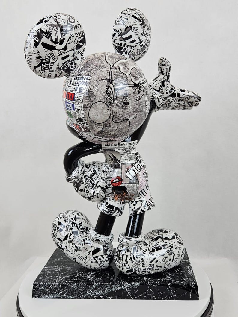 Pode incluir: Uma figura de Mickey Mouse em preto e branco, coberta de recortes de jornais e texto. A figura est&aacute; em p&eacute; sobre uma base de m&aacute;rmore preto e branco.
