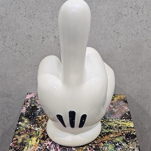 Pode incluir: Uma escultura de cerâmica branca de uma mão a fazer o gesto do dedo do meio. A mão tem detalhes pretos na palma e está colocada numa superfície colorida com um padrão abstrato. É uma obra de arte moderna.