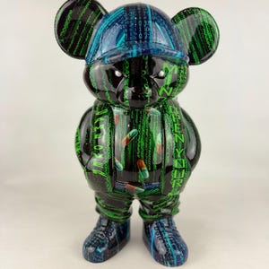 Peut inclure: Une figurine en forme d'ours avec une base noire et un motif de code numérique matriciel vert. La figurine porte une casquette et des vêtements ornés du texte "GAME OVER" et "OPEN YOUR MIND", ainsi que de petits graphismes de pilules rouges et vertes.