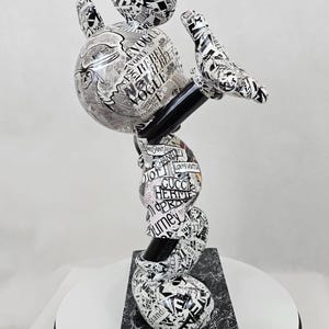 Pode incluir: Uma escultura em preto e branco do Mickey Mouse, feita de papel e coberta de recortes de revistas. A escultura est&aacute; em uma pose din&acirc;mica, quase acrob&aacute;tica, com os bra&ccedil;os e as pernas do Mickey estendidos. Os recortes de revistas apresentam marcas e logotipos de moda, incluindo Vogue, Chanel, Gucci e Louis Vuitton.