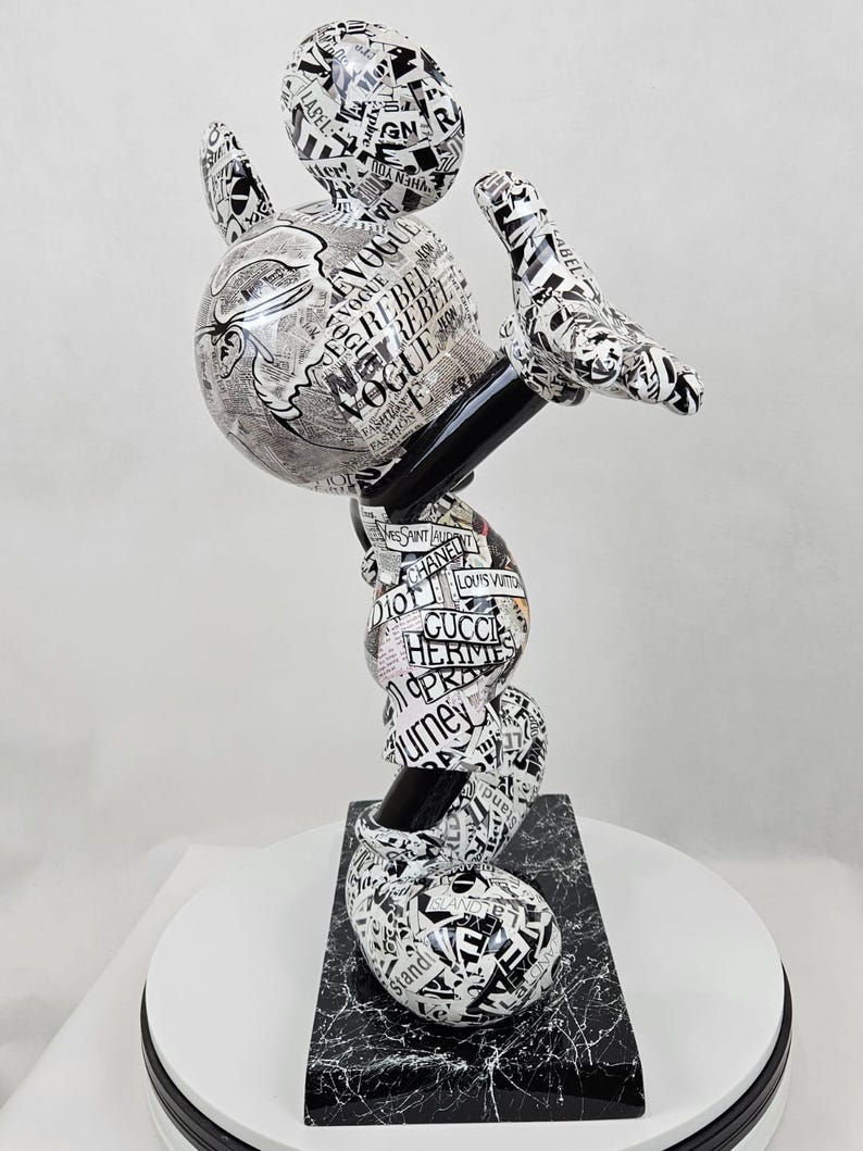 Pode incluir: Uma escultura em preto e branco do Mickey Mouse, feita de papel e coberta de recortes de revistas. A escultura est&aacute; em uma pose din&acirc;mica, quase acrob&aacute;tica, com os bra&ccedil;os e as pernas do Mickey estendidos. Os recortes de revistas apresentam marcas e logotipos de moda, incluindo Vogue, Chanel, Gucci e Louis Vuitton.
